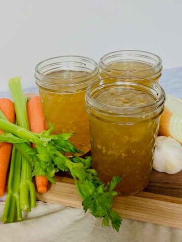 mason jars of chicken broth next to carrot celery and onion on a wood cutting board