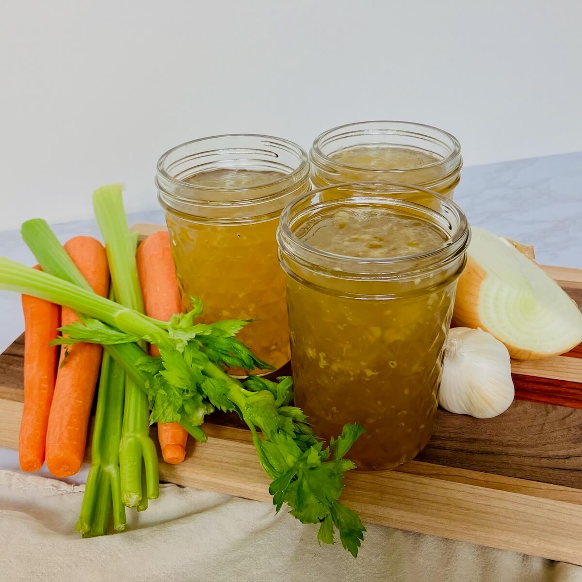 mason jars of chicken broth next to carrot celery and onion on a wood cutting board