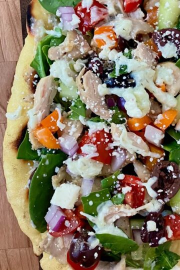 close up of a greek chicken flatbread with all of the veggie and tzatziki toppings on a wooden cutting board