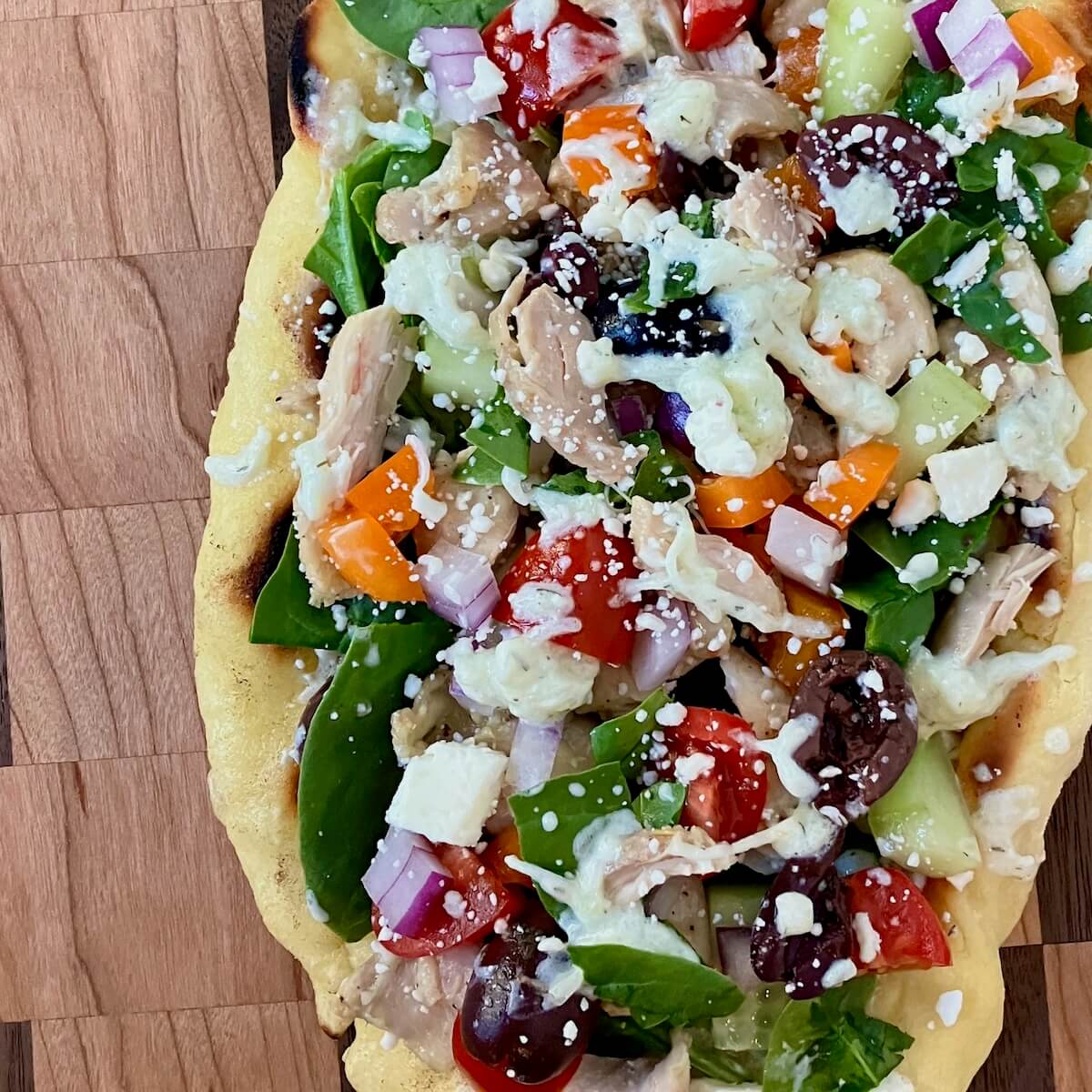 close up of a greek chicken flatbread with all of the veggie and tzatziki toppings on a wooden cutting board