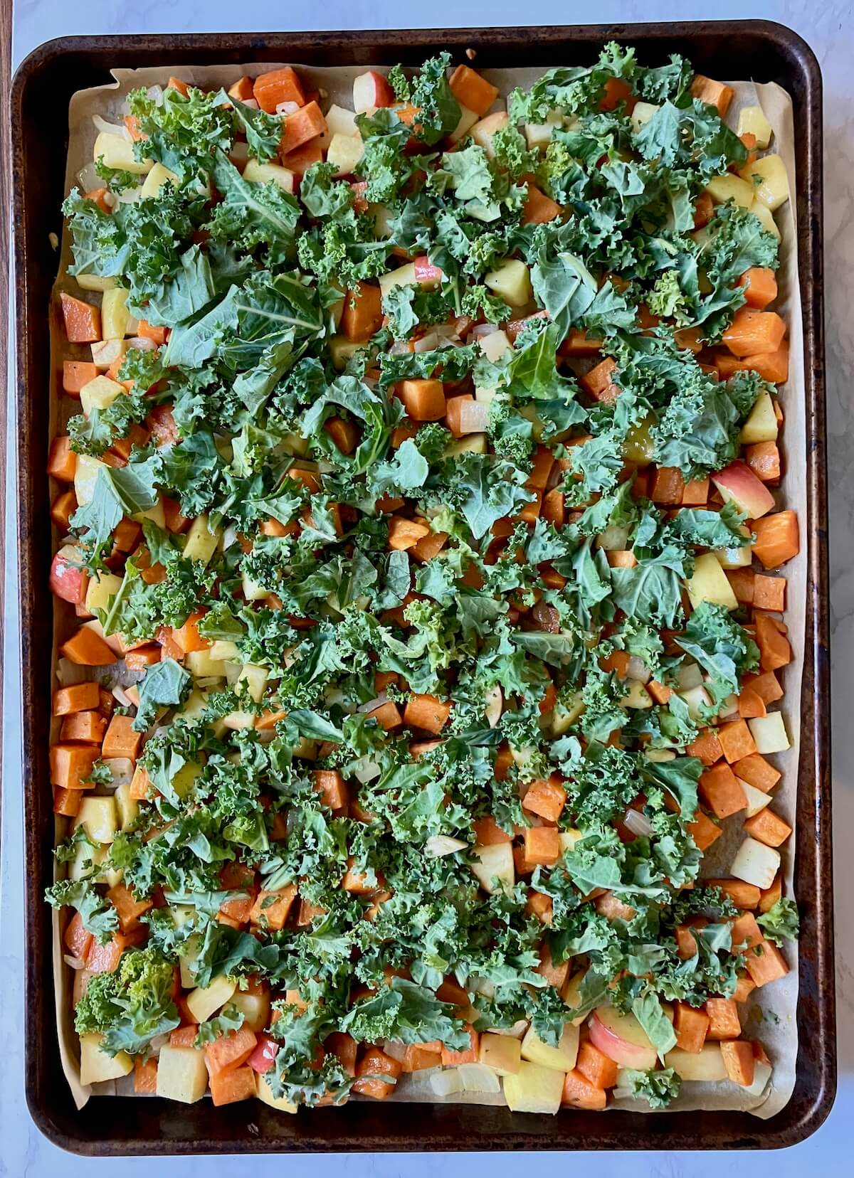 cooked cubed sweet potato, apple and onion on a baking sheet topped with a layer of kale