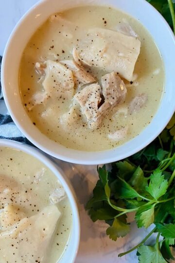 two white bowls of chicken and dumplings on a white background with green herbs