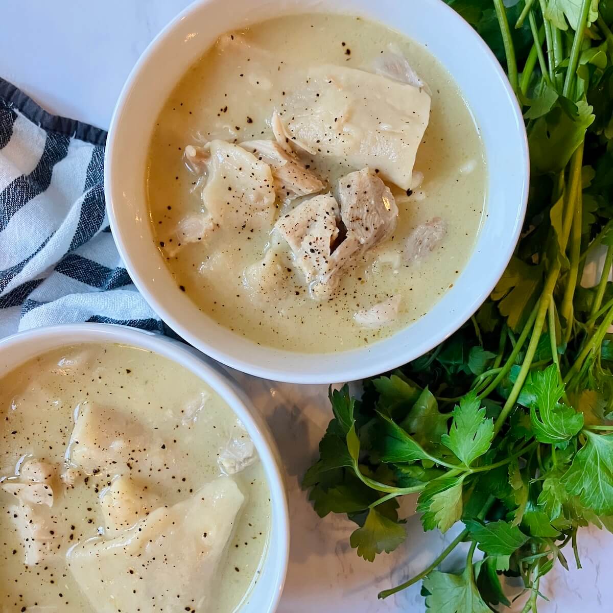 two white bowls of chicken and dumplings on a white background with green herbs