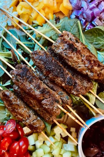 beef koftas on skewers on top of lettuce surrounded by chopped veggies, feta and olives