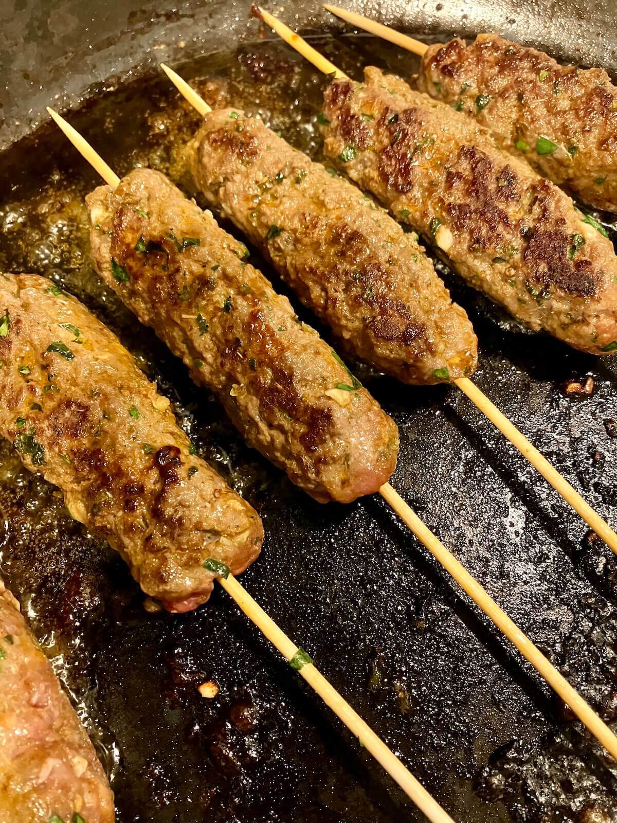 beef koftas on skewers cooking on a dark pan